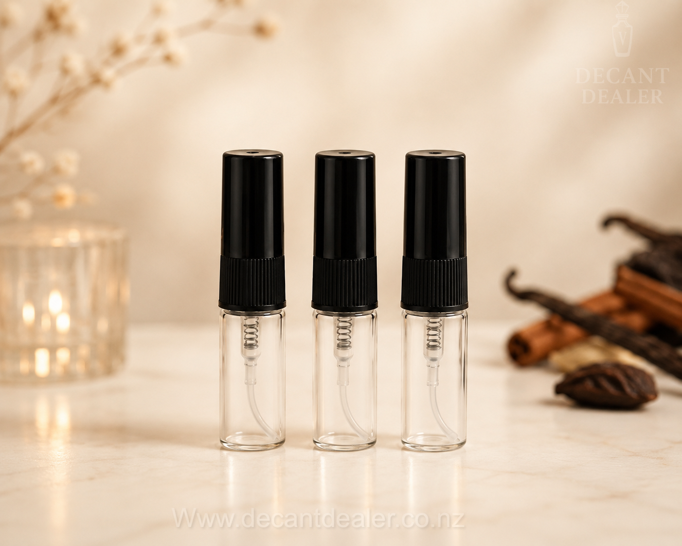 Three small clear glass spray bottles with black atomizers standing on a light marble surface, with warm decorative props and dried flowers in the background. A faint 'DECANT DEALER' watermark appears in the top right, and a website watermark is at the bottom.