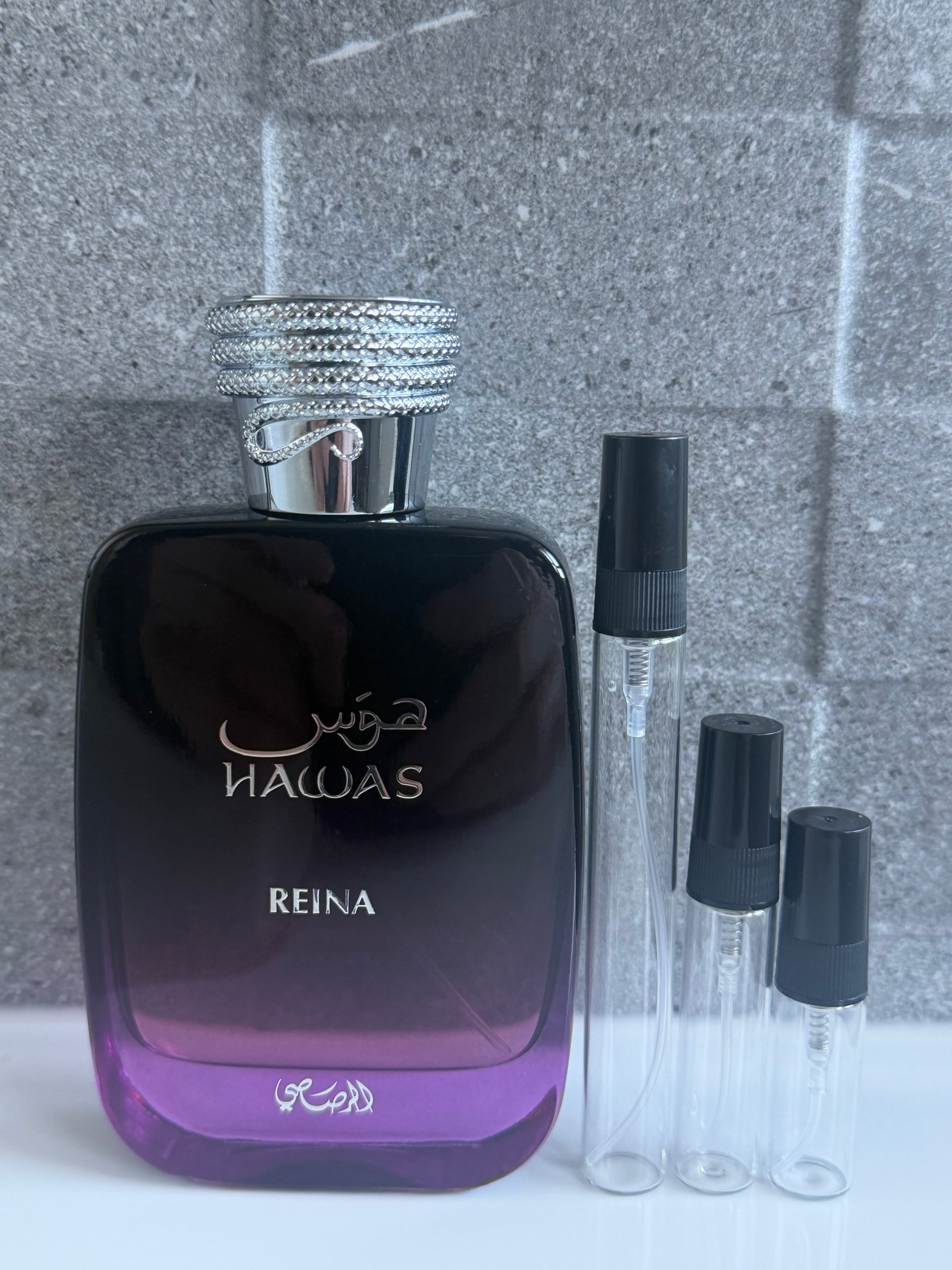 A bottle of Hawas Reina perfume with a dark purple-to-black gradient body, silver cap, and three clear sample spray vials with black caps, set against a gray tiled background. Decant sizes next to it3ml, 5ml and 10ml.