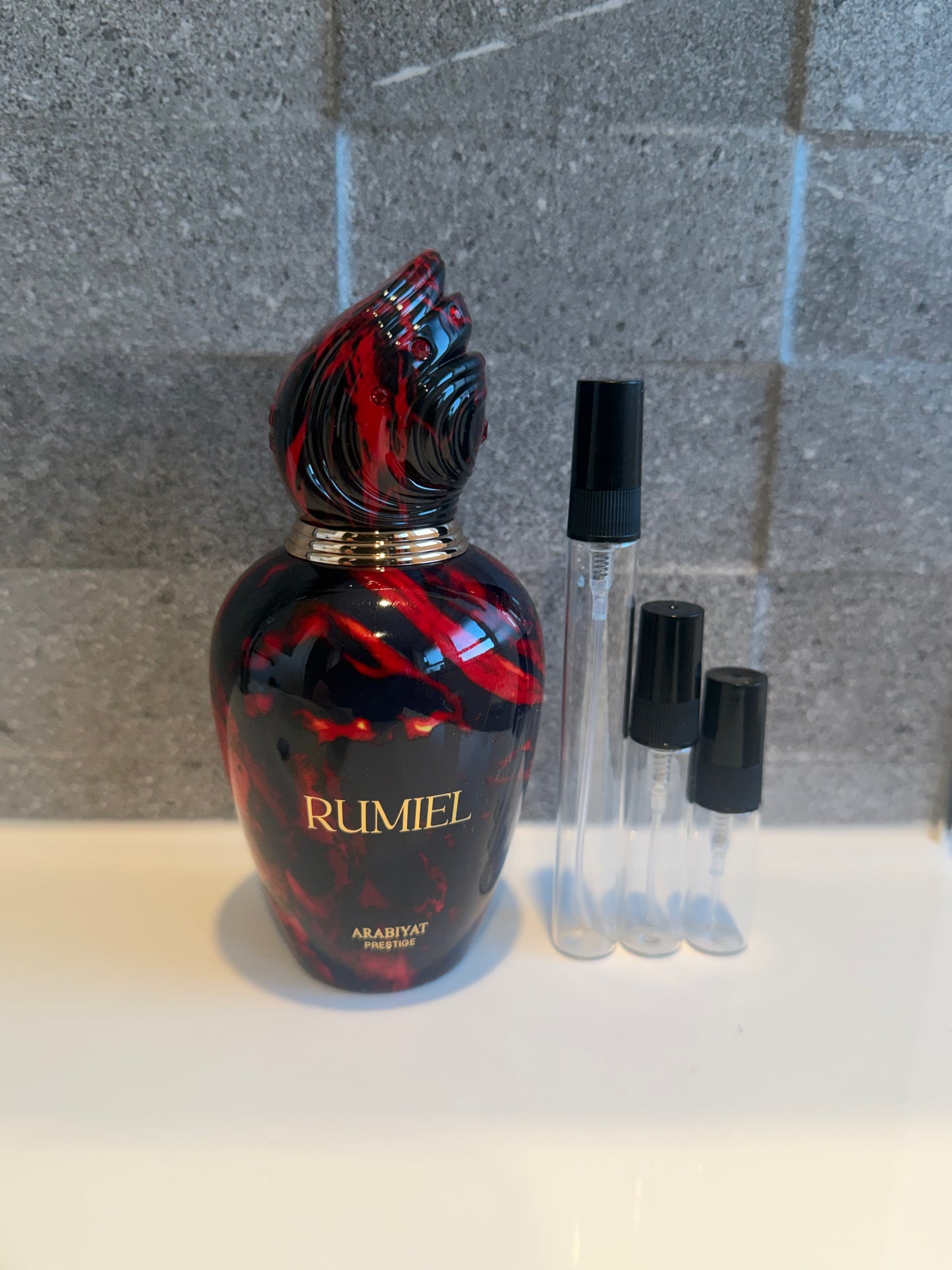 A bottle of Rumiel Arabian perfume with a black and red marbled design and gold lettering, displayed next to three clear sample spray vials with black caps on a white surface against a gray tiled background. Decant Dealer nz decant sizes 