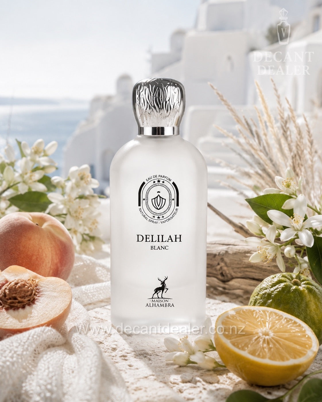 A frosted glass bottle of Delilah Blanc liqueur by maison alhrambra, styled in a Mediterranean setting with white buildings in the background, surrounded by peaches, a halved lemon, a lime, and white blossoms.