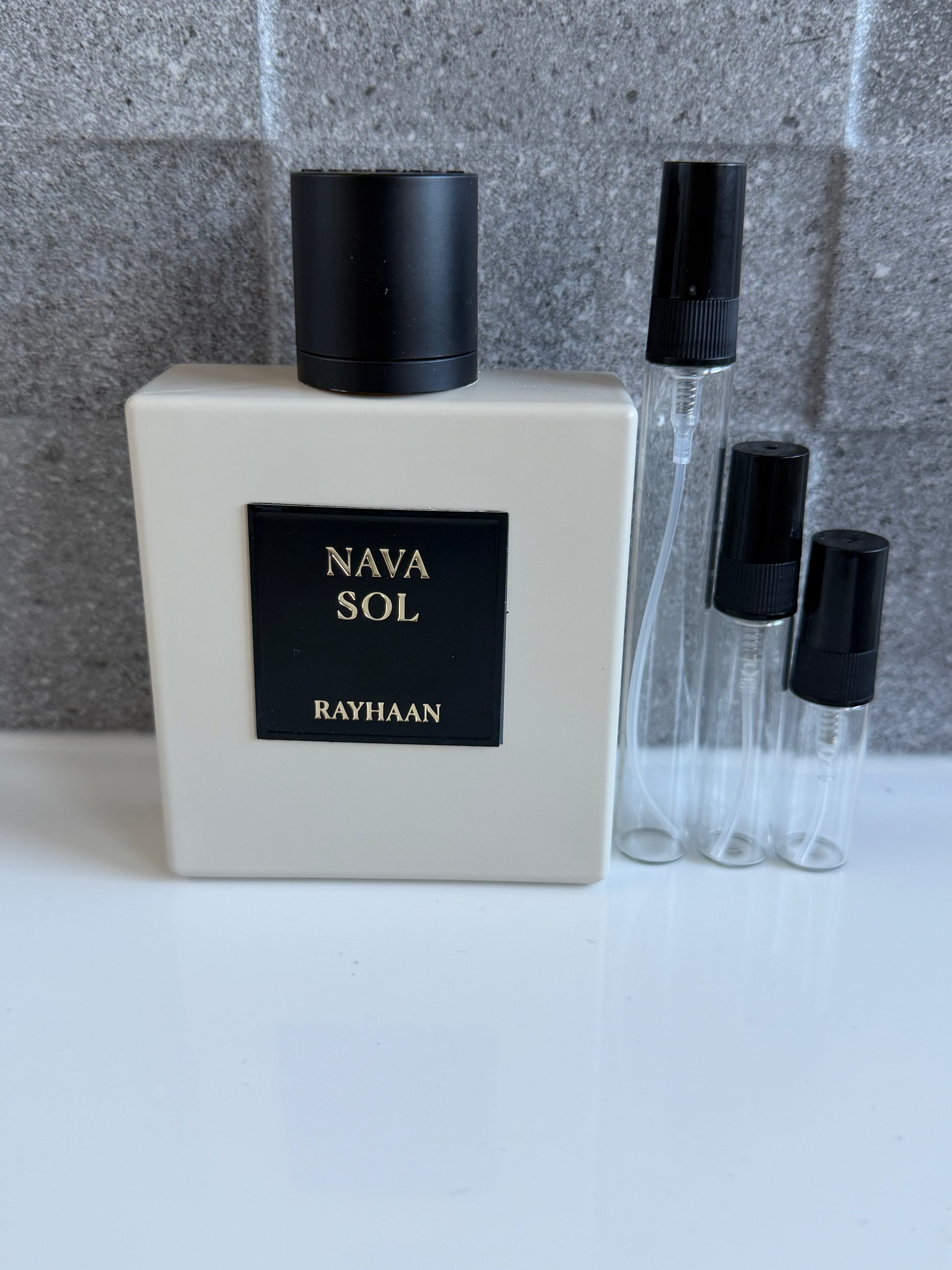 A bottle of Nava Sol fragrance by Rayhaan with three clear sample spray vials, set against a gray tiled background on a white surface.