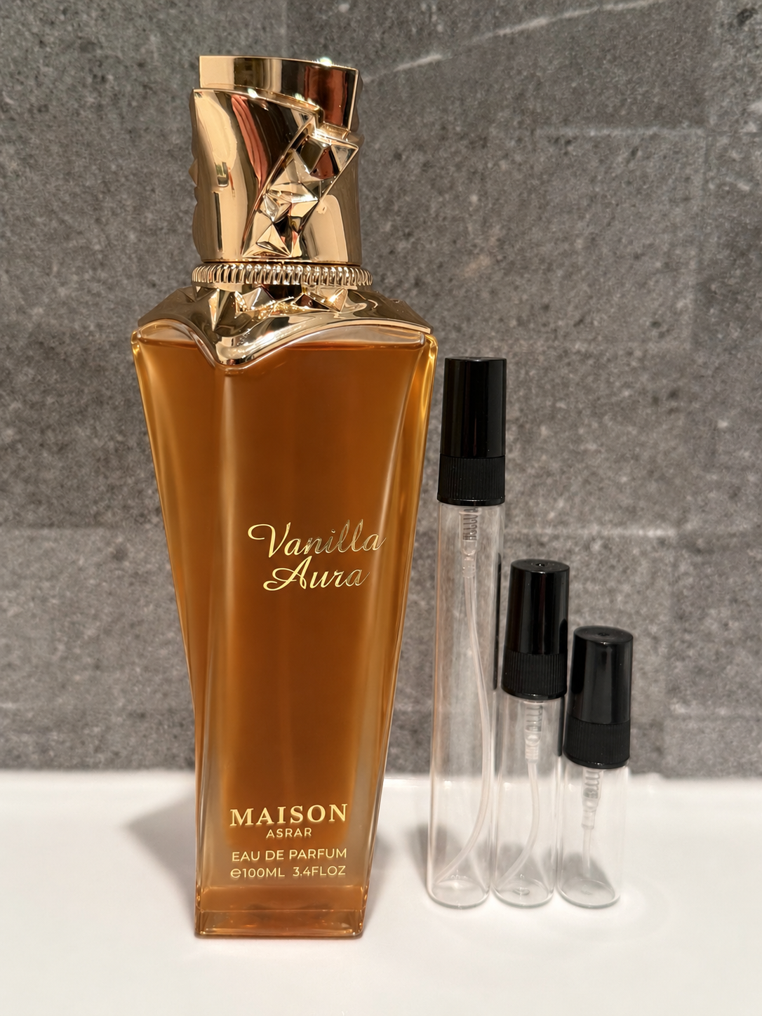 Maison Asra Vanilla Aura Eau de Parfum bottle in amber glass with a metallic gold cap, shown next to three clear sample spray vials with black caps on a white surface against a gray textured background. Decant Dealer nz 